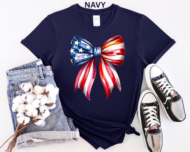 May include: Navy blue t-shirt featuring a watercolor bow design in red, white, and blue, with stars and stripes. The word "NAVY" is printed at the top. The shirt is styled with denim shorts and black sneakers.