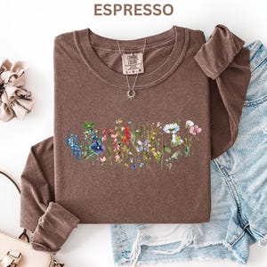 May include: Espresso-colored long-sleeve shirt with a floral design across the front. The shirt has a crew neck and is paired with denim shorts. A necklace with a sun pendant and a scrunchie are also visible.
