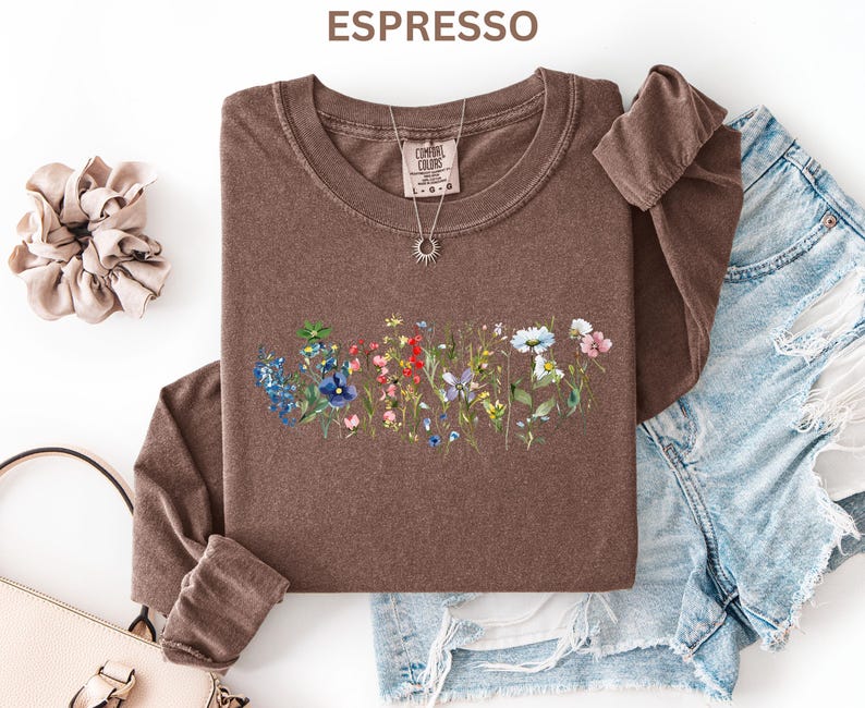 May include: Espresso-colored long-sleeve shirt with a floral design across the front. The shirt has a crew neck and is paired with denim shorts. A necklace with a sun pendant and a scrunchie are also visible.