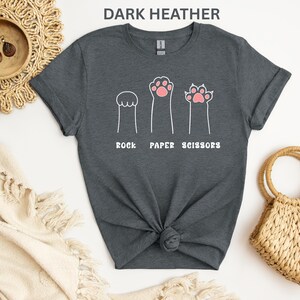 Puede incluir: Camiseta gris oscuro con un divertido dise&ntilde;o de patas de gato que representan piedra, papel y tijeras. El texto blanco "ROCK PAPER SCISSORS" est&aacute; impreso debajo. Una prenda casual para el d&iacute;a a d&iacute;a.