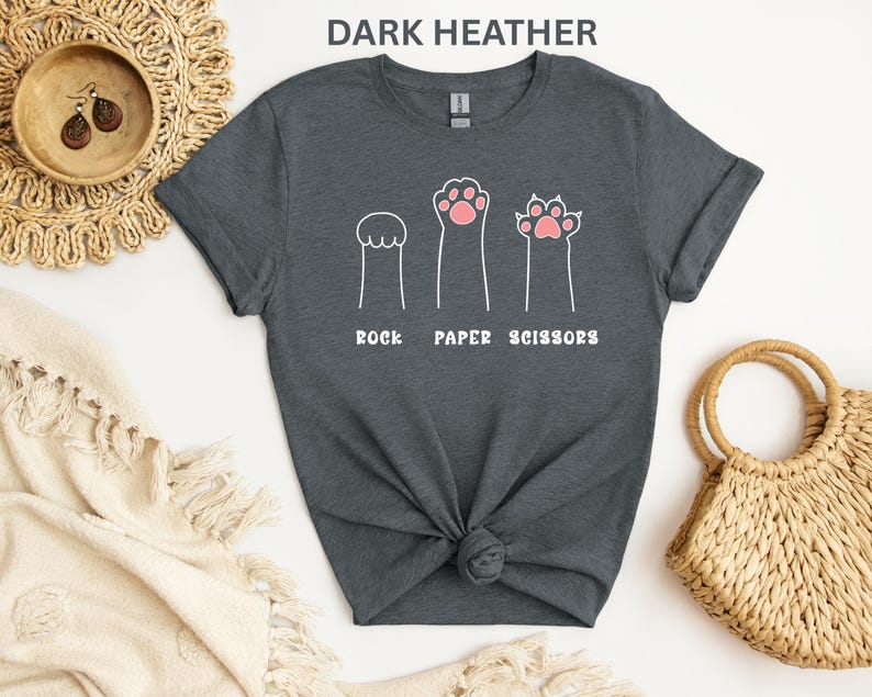 Puede incluir: Camiseta gris oscuro con un divertido dise&ntilde;o de patas de gato que representan piedra, papel y tijeras. El texto blanco "ROCK PAPER SCISSORS" est&aacute; impreso debajo. Una prenda casual para el d&iacute;a a d&iacute;a.
