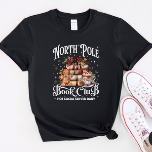 May include: A black short-sleeve t-shirt featuring a graphic with the text North Pole Book Club and Hot Cocoa Served Daily. The design includes a stack of books, a gingerbread person, and a mug with the word Read, accented by holly and stars.