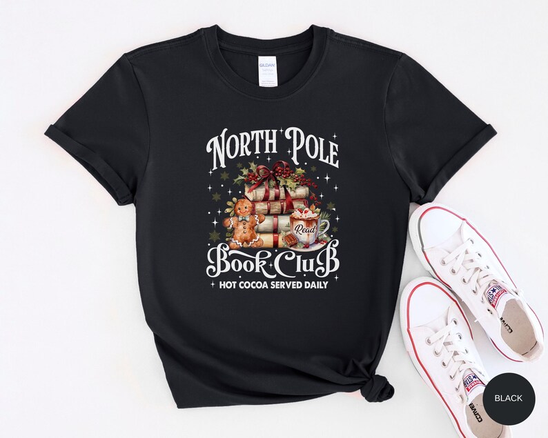 May include: A black short-sleeve t-shirt featuring a graphic with the text North Pole Book Club and Hot Cocoa Served Daily. The design includes a stack of books, a gingerbread person, and a mug with the word Read, accented by holly and stars.