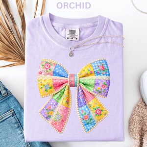 May include: A lilac t-shirt with a patchwork bow design. The bow is made up of floral patterns in pink, yellow, blue, and green. The word "ORCHID" is at the top. A silver necklace and blue jeans are also visible.