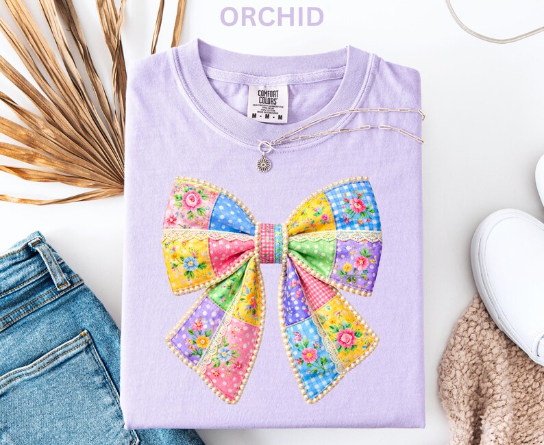 May include: A lilac t-shirt with a patchwork bow design. The bow is made up of floral patterns in pink, yellow, blue, and green. The word "ORCHID" is at the top. A silver necklace and blue jeans are also visible.