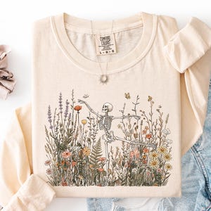 Puede incluir: Una camiseta de manga larga de color crema con un gr&aacute;fico de un esqueleto bailando en medio de un campo de flores silvestres coloridas y mariposas. La camiseta tiene cuello redondo y una etiqueta de la marca Comfort Colors en el interior.