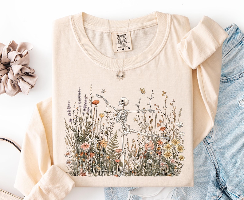 Puede incluir: Una camiseta de manga larga de color crema con un gr&aacute;fico de un esqueleto bailando en medio de un campo de flores silvestres coloridas y mariposas. La camiseta tiene cuello redondo y una etiqueta de la marca Comfort Colors en el interior.