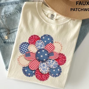 May include: Cream-colored t-shirt with a faux patchwork flower design. The flower features red, white, and blue patterned circles, including American flags, stars, and gingham patterns. The text "FAUX PATCHWORK" is visible.