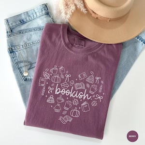 May include: A berry-colored t-shirt featuring a white line-art heart design composed of small icons including books, pumpkins, mugs, and mittens, with the word bookish centered in the heart, paired with light blue denim jeans and a tan hat.
