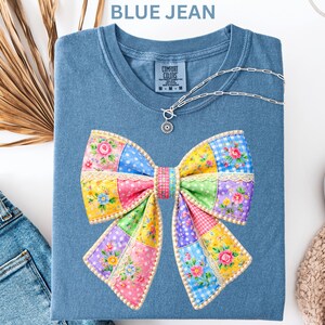 May include: A blue jean t-shirt with a patchwork bow design. The bow is made up of various patterns and colours, including pink, yellow, blue, and green. A silver necklace with a sun pendant is on the shirt. The text "BLUE JEAN" is at the top.