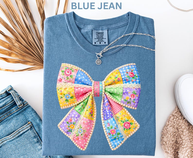 May include: A blue jean t-shirt with a patchwork bow design. The bow is made up of various patterns and colours, including pink, yellow, blue, and green. A silver necklace with a sun pendant is on the shirt. The text "BLUE JEAN" is at the top.
