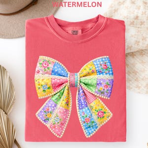 May include: A coral-coloured t-shirt with a large patchwork bow design. The bow features various patterns, including floral prints, polka dots, and gingham, in shades of pink, blue, yellow, and green. The word "WATERMELON" is printed at the top.