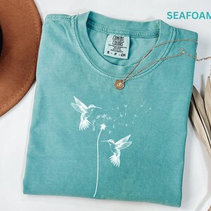 a turquoise t-shirt with a white bird design, a brown hat, and a necklace, all laid out on a white surface.