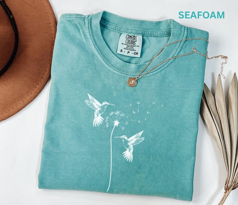 a turquoise t-shirt with a white bird design, a brown hat, and a necklace, all laid out on a white surface.