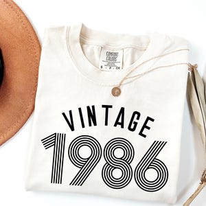 May include: Cream-colored t-shirt with the word "VINTAGE" above the number "1986" in black. A gold necklace with a pendant is draped over the shirt. A brown hat and dried plant are in the background.