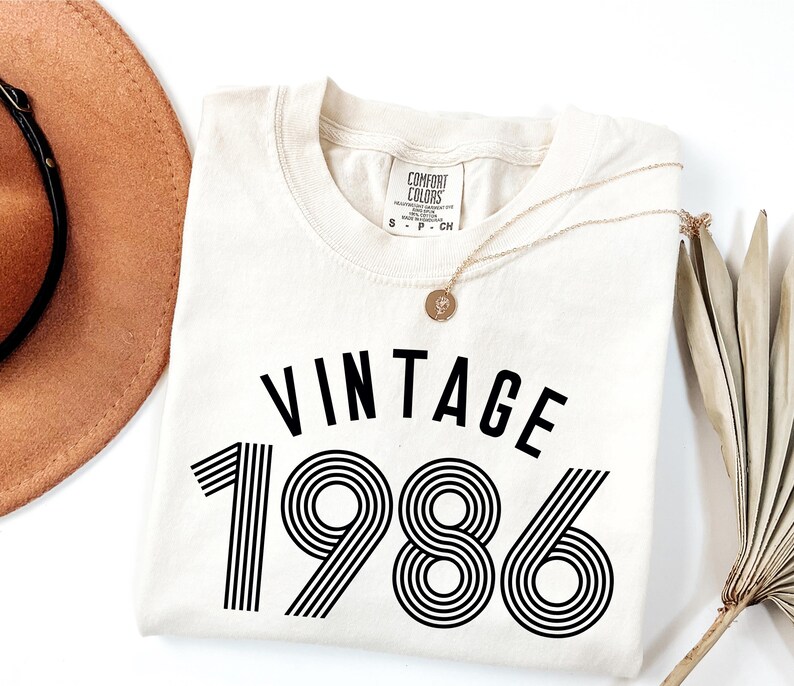 May include: Cream-colored t-shirt with the word "VINTAGE" above the number "1986" in black. A gold necklace with a pendant is draped over the shirt. A brown hat and dried plant are in the background.