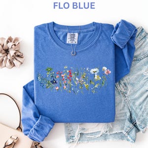 May include: A blue long-sleeved shirt with a floral design across the chest. The shirt is paired with denim shorts, a scrunchie, and a purse. The text "FLO BLUE" is at the top of the image. A silver necklace is on the shirt.