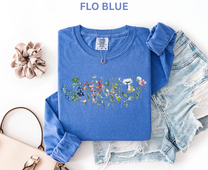 May include: A blue long-sleeved shirt with a floral design across the chest. The shirt is paired with denim shorts, a scrunchie, and a purse. The text "FLO BLUE" is at the top of the image. A silver necklace is on the shirt.
