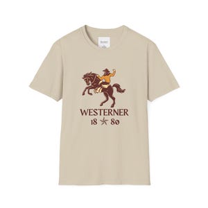 May include: A tan-colored t-shirt featuring a graphic of a cowboy riding a horse. The text "WESTERNER 18 80" is printed below the image in a brown color. The shirt has a crew neck and short sleeves.