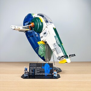 Op de afbeelding: Een Lego Star Wars model van Jango Fett's Starship, met blauwe, groene, witte en gele kleuren. Het model wordt gepresenteerd op een transparante standaard met een zwarte basis, met een informatiekaart en twee Lego-figuren.