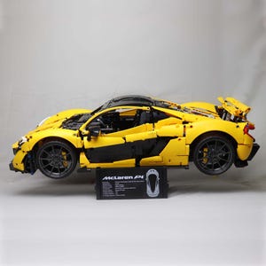 May include: A yellow Lego McLaren P1 model car with black accents and a black base. The car is on a white surface.