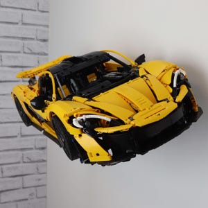 May include: A yellow and black Lego McLaren 720S model car. The car is built with Lego bricks and is displayed on a white wall.