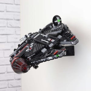 Può includere: Un modello nero e grigio di un'astronave fatto con mattoncini, montato su un muro bianco. Il modello presenta dettagli rossi e verdi, con una cupola rossa trasparente nella parte anteriore. L'astronave è angolata, mostrando il suo design dettagliato.