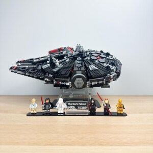 Peut inclure: Maquette Lego noire du vaisseau spatial Dark Falcon, avec des accents rouges et gris, exposée sur un support transparent. Comprend six figurines Lego sur une base noire. La base porte l'inscription "The Dark Falcon 75389".
