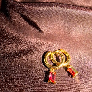 Peut inclure: Une paire de boucles d'oreilles créoles dorées avec des pendentifs rectangulaires en pierres précieuses rouges. Les boucles d'oreilles sont présentées sur un tissu de couleur marron foncé, satiné. Les pierres sont d'un rouge vif.