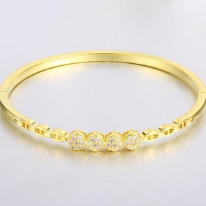 May include: A gold-coloured bangle bracelet featuring four oval-shaped, diamond-like embellishments. The bracelet has a simple, elegant design, suitable for everyday wear or special events. The bracelet is made of a shiny, reflective metal.