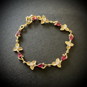 Könnte beinhalten: Ein goldenes Armband mit einem zarten Design aus roten und weißen Edelsteinen. Das Armband besteht aus einer Reihe kleiner Blätter mit einem tropfenförmigen Edelstein in der Mitte jedes Blattes.