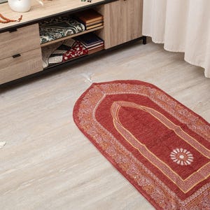 May include: A red prayer mat with an elaborate border and a central floral pattern is shown on a light wood floor. A wooden cabinet holds books, prayer beads, and a decorative Quran stand. A white vase with dried plants is on the cabinet.