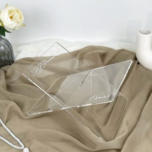 May include: A clear acrylic book stand with a decorative design and the name "Omar" engraved on it. The stand is sitting on a beige fabric surface.