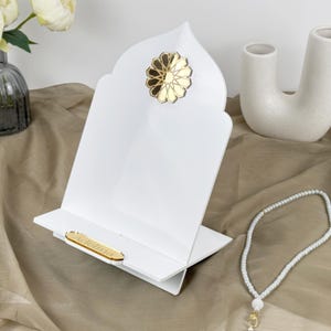 Könnte beinhalten: Ein weißer Holzbuchständer mit einem goldenen Dekor und einer goldenen Inschrift, die "Alhamdulillah" lautet. Der Ständer steht auf einer braunen Stoffoberfläche mit einer weißen Gebetsperlenkette und einem weißen Quasten.