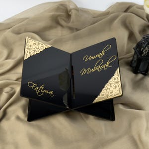 Pode incluir: Suporte para livros de acrílico preto com detalhes dourados e as palavras "Fatima" e "Umrah Mubarak" escritas em caligrafia dourada.