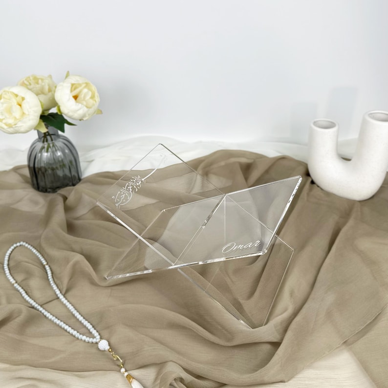 May include: A clear acrylic book stand with a decorative design and the name "Omar" engraved on it. The stand is sitting on a beige fabric surface.