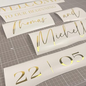 May include: Gold foil lettering on white vinyl, including the words "WELCOME TO OUR BEGINNING", "Thomas", "Michelle", "and", and the numbers "22 05". The pieces are arranged on a gray cutting mat.