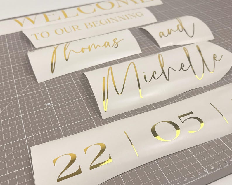 May include: Gold foil lettering on white vinyl, including the words "WELCOME TO OUR BEGINNING", "Thomas", "Michelle", "and", and the numbers "22 05". The pieces are arranged on a gray cutting mat.
