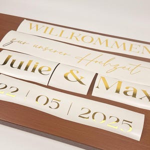 May include: Several white vinyl decals with gold lettering are arranged on a wooden surface. The decals include phrases like "Willkommen", "Julie & Max", and a date. The overall theme is wedding decor.