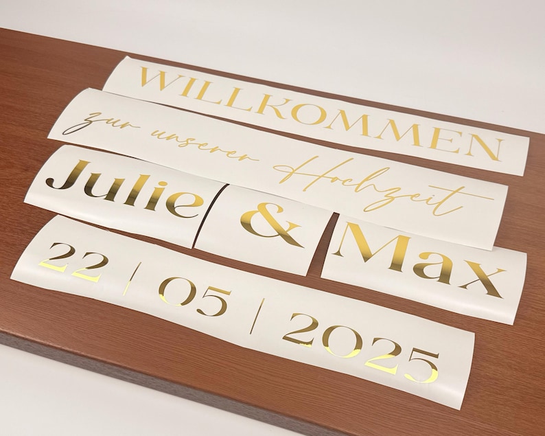 May include: Several white vinyl decals with gold lettering are arranged on a wooden surface. The decals include phrases like "Willkommen", "Julie & Max", and a date. The overall theme is wedding decor.