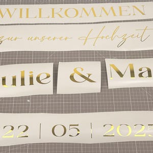 May include: Gold foil lettering on white vinyl. The text includes "WILLKOMMEN", "zur unserer Hochzeit", "Julie & Max", and the date "22 | 05 | 2025". The lettering is for a wedding.