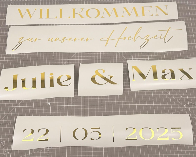 May include: Gold foil lettering on white vinyl. The text includes "WILLKOMMEN", "zur unserer Hochzeit", "Julie & Max", and the date "22 | 05 | 2025". The lettering is for a wedding.