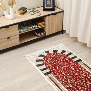 Op de afbeelding: Een rood gebedskleed met een bloemmotief en een decoratief boogontwerp. Het kleed ligt op een lichte houten vloer. Een houten kast met boeken en een decoratieve Koranstandaard is op de achtergrond te zien.
