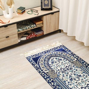 May include: A blue and white prayer rug with a floral design and fringe. The rug is placed on a light wood floor. A wooden cabinet with books and a decorative frame is in the background.