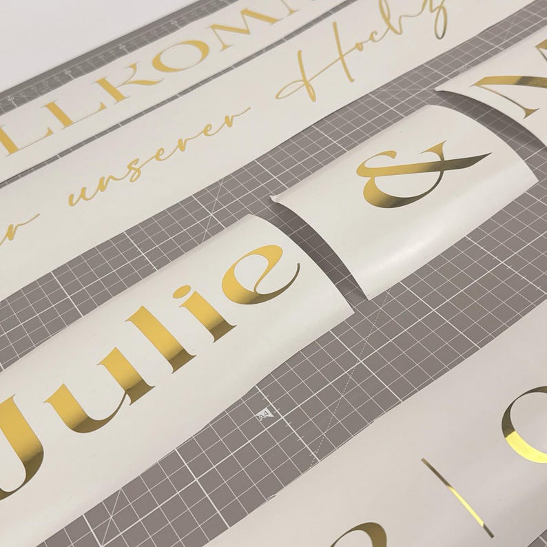 May include: Gold foil lettering on white vinyl sheets. The image shows various phrases and names, including "Julie" and "&", likely for wedding or event decorations. The gold lettering contrasts with the white background, creating a visually appealing design.