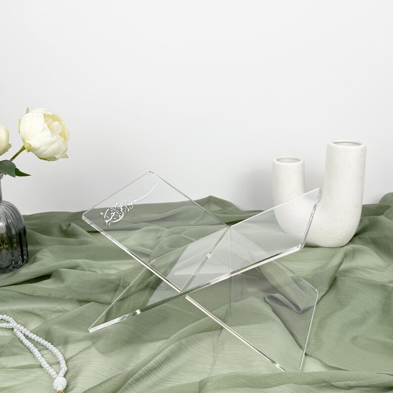 May include: A clear acrylic book stand with an etched design. The stand is folded and resting on a green fabric surface.