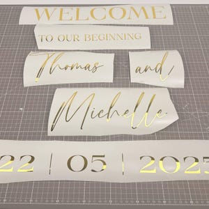 May include: Gold lettering on white vinyl pieces. Text includes "WELCOME", "TO OUR BEGINNING", names "Thomas and Michelle", and the date "22|05|2025". Ideal for wedding or event decor.