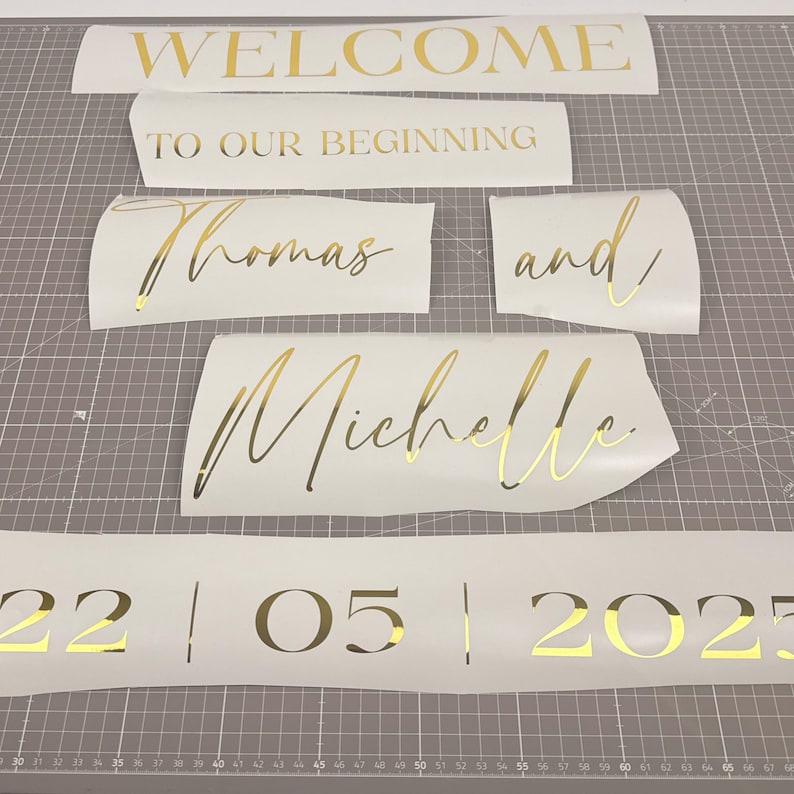 May include: Gold lettering on white vinyl pieces. Text includes "WELCOME", "TO OUR BEGINNING", names "Thomas and Michelle", and the date "22|05|2025". Ideal for wedding or event decor.