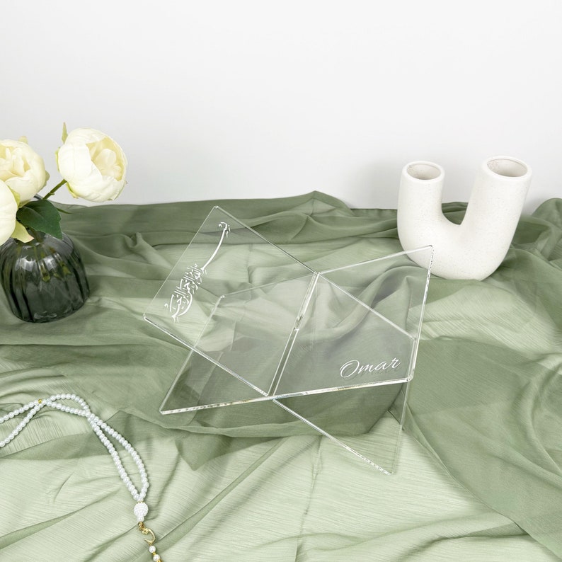 May include: A clear acrylic book stand with a decorative design and the name "Omar" engraved on it. The stand is folded and sitting on a green fabric surface.
