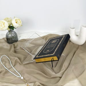 May include: A clear acrylic book stand with a black book titled "Al-Quran Al-Karim" resting on top. The book stand has a decorative inscription in Arabic.